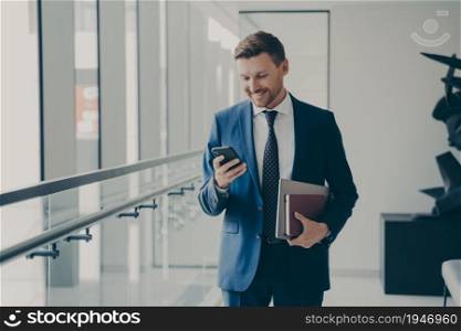 Happy handsome man ceo executive holding mobile phone and smiling, reading good news from business partner while standing in office with laptop in hand, chatting with friend during coffee break. Happy handsome man ceo executive holding mobile phone and smiling during coffee break in office
