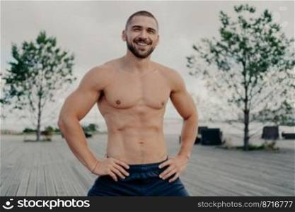 Happy handsome man with thick bristle keeps hands on waist, stands with naked muscular torso poses outdoor breathes fresh air. Athletic male looks positively into distance. Sport workout, bodybuilding