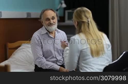 Happy handsome senior male patient with beard receiving good news from female physician in hospital office. Caring woman doctor talking with elderly man and explaining the good medical results to patient in clinic, who is happy and relieved..