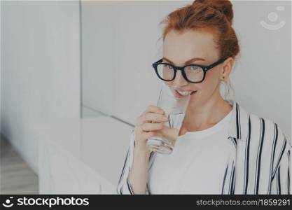 Happy healthy beautiful young redhead woman with clean skin drinking water in morning and smiling, beautiful female holding glass in hand, preventing dehydration while standing in kitchen at home. Happy healthy beautiful young redhead woman drinking water in morning at home