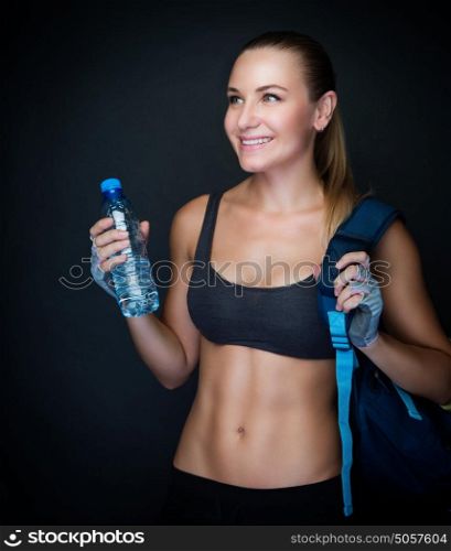 Happy healthy sportive woman over dark background, fitness trainer wearing sexy outfit and holding bottle of water, healthy lifestyle