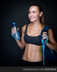 Happy healthy sportive woman over dark background, fitness trainer wearing sexy outfit and holding bottle of water, healthy lifestyle