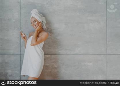 Happy healthy young woman with tender facial skin, applies beauty cream and undergoes beauty treatments, wrapped in bath towel, poses against grey background with copy space for your promotion