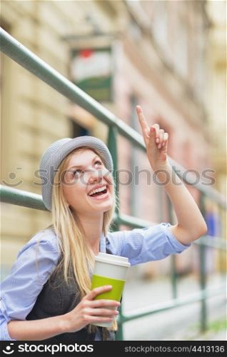 Happy hipster girl with cup of hot beverage pointing on copy space on city street