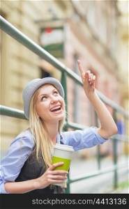 Happy hipster girl with cup of hot beverage pointing on copy space on city street