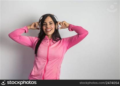 Happy Hispanic female in activewear keeping hands on wireless headphones and enjoying songs for training while looking at camera against white background. Smiling ethnic woman listening to playlist in headphones