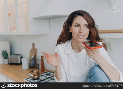 Happy hispanic female sends audio voice message by smartphone in kitchen at home. Modern woman chatting on social networks, uses speaker phone, virtual assistant to search information on internet.. Happy hispanic female sends audio voice message, chatting, using speaker phone in kitchen at home