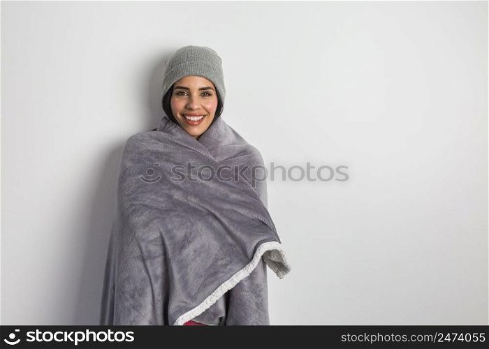 Happy Hispanic female warming with soft blanket and in beanie while standing against white background and looking at camera. Smiling ethnic woman in warm blanket and hat