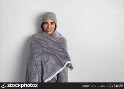 Happy Hispanic female warming with soft blanket and in beanie while standing against white background and looking at camera. Smiling ethnic woman in warm blanket and hat