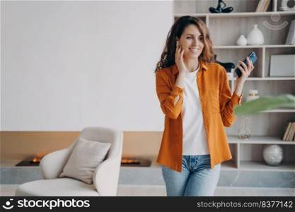 Happy hispanic girl has phone call at home. Young woman in casual wear is using wireless earphones. Headset using. Technology, leisure and communication concept. Beautiful modern interior.. Happy girl has phone call at home using wireless earphones. Technology, leisure and communication.