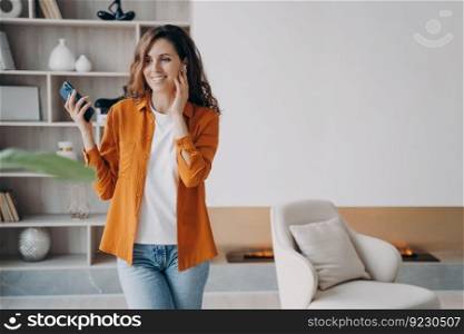 Happy hispanic girl has phone call at home. Young woman in casual wear is using wireless earphones. Headset using. Technology, leisure and communication concept. Beautiful modern interior.. Happy girl has phone call at home using wireless earphones. Technology, leisure and communication.