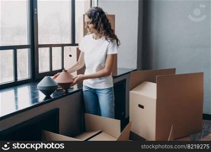 Happy hispanic girl unpacks carton box with things at home, puts vases on window sill. Woman unpacking belongings, relocating at new house. Female received parcel from transport service on moving day.. Happy girl unpacks carton box with things at new home on moving day. Transport service advertising