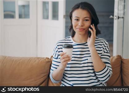 Happy hispanic woman has conversation on mobile phone. Lady is sitting on couch enjoying cup of tea and smiling. Woman in striped sailor style shirt. Remote work online at cozy home.. Happy lady sitting on couch enjoying cup of tea and smiling. Woman has conversation on mobile phone.