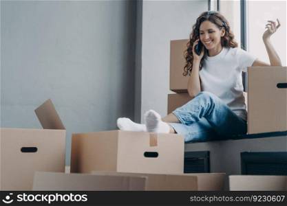 Happy hispanic woman is sitting on windowsill of new house. Girl is unpacking boxes and talking on phone to friends. Relocation and communication concept.. Happy hispanic woman sitting on windowsill of new house. Girl unpacking boxes and talking on phone.