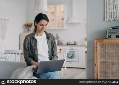 Happy hispanic woman using laptop sitting on sofa in living room at home. Smiling female typing email, working online, learning language remotely. Distant work, remote education concept.. Female freelancer working online using laptop sitting on sofa in living room at home. Distant work