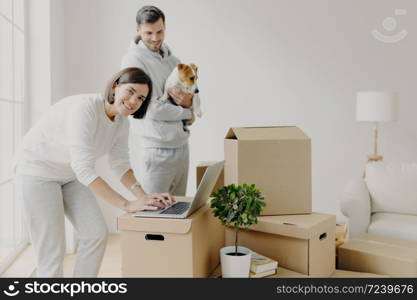 Happy home buyers pose near unpacked boxes, enjoy relocation in new house, woman searches ideas for redecoration bedroom, man carries pedigree dog, decide to do repairing of flat. Mortgage concept