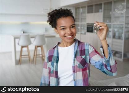 Happy homeowner mixed race girl celebrating relocation to new home, showing key to first own flat. Smiling young lady holding keys, standing in modern apartment studio, looking at camera.. Happy young mixed race girl celebrating relocation to new home, showing key to first own apartment