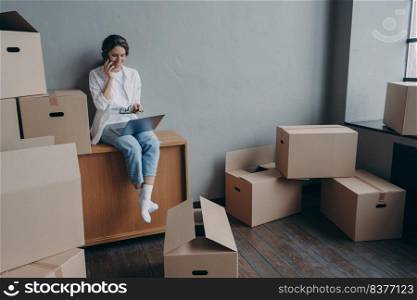 Happy homeowner talking on phone. Hispanic girl is sitting among carton boxes and working on pc. Attractive young business woman moves. Mortgage loan and real estate purchase.. Happy homeowner talking on phone. Hispanic girl is sitting among carton boxes and working on pc.