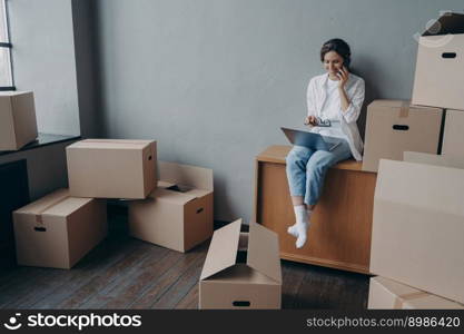 Happy homeowner talking on phone. Hispanic girl is sitting among carton boxes and working on pc. Attractive young business woman moves. Mortgage loan and real estate purchase.. Happy homeowner talking on phone. Hispanic girl is sitting among carton boxes and working on pc.