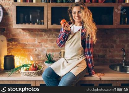 Happy housewife in apron sits on kitchen table and holds tomato in hand. Female cook making healthy vegetarian food, vegetables preparation. Housewife sits on table and holds tomato in hand
