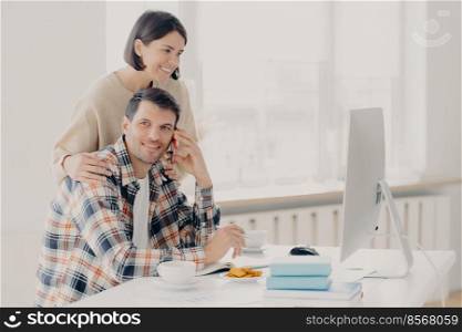 Happy husband and wife do paperwork together, pay taxes online look at screen computer, pose at desktop with books and papers, drink coffee and have lunch, make telephone call via smartphone