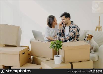 Happy husband and wife move in new bought house, buy furniture online, use modern technology, man surfs internet via laptop and talk over phone, sits on sofa with girlfriend and dog, many carton boxes