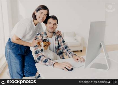 Happy husband and wife pose near computer, keyboard and surf information, work in domestic atmosphere with dog, pose in coworking space, connected to wireless internet. Lovely couple at desktop