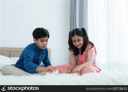 Happy Indian family, brother and sister with traditional clothes playing fun game together, teenage boy and girl sitting on bed at home, sibling relationship concept