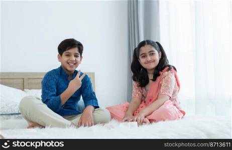 Happy Indian family, brother and sister with traditional clothes smiling and looking at camera while playing fun game, sitting together on bed at home, sibling relationship concept