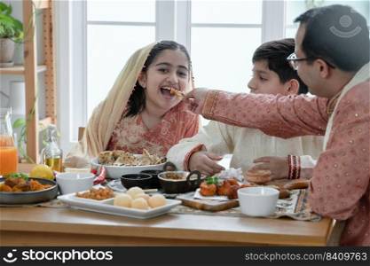 Happy Indian family enjoy eating food with hands, se≤ctive focus on Asian cute girl eating naan bread dipπng in curry from father feeding, wear traditional clothes, sitting with brother at home