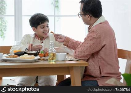 Happy Indian family enjoy eating food with hands, selective focus on South Asian cute boy eating naan bread dipping in curry from father feeding, wear traditional clothes, sitting at dining table