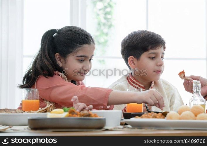 Happy Indian family enjoy eating food with hands, selective focus on South Asian cute girl holding naan bread dipping in curry, wear traditional clothes, sitting with brother at dining table at home