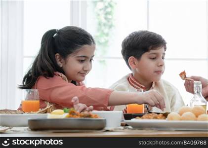Happy Indian family enjoy eating food with hands, selective focus on South Asian cute girl holding naan bread dipping in curry, wear traditional clothes, sitting with brother at dining table at home