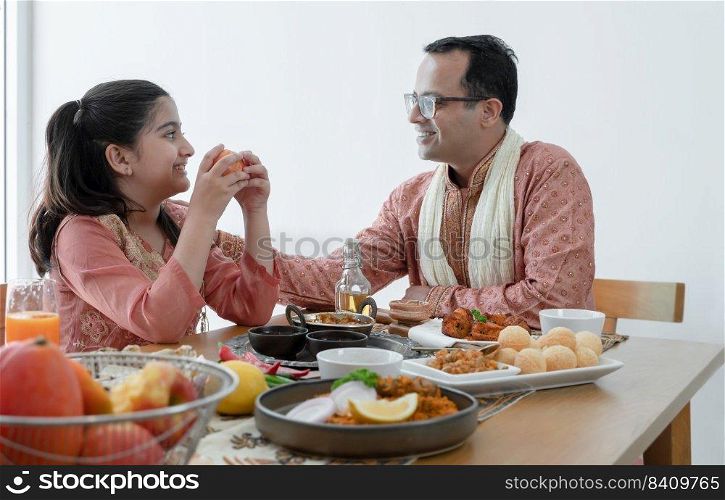 Happy Indian family enjoy eating food with hands, South Asian father and cute daughter eating apple after meal, wearing traditional clothes, sitting and talking at dining table at home together