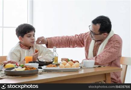 Happy Indian family enjoy eating food with hands, South Asian father wipe his little boy mouth with love and care, wear traditional clothes, sitting and talking at dining table at home together