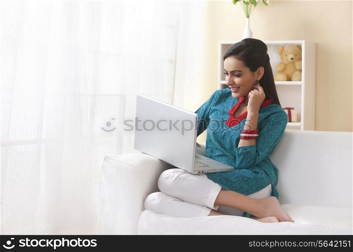 Happy Indian woman using laptop on sofa