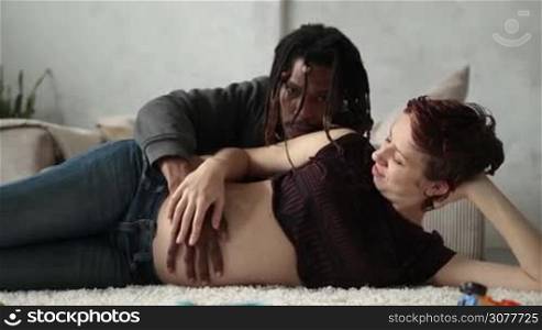 Happy interracial couple expecting baby. Smiling caucasian pregnant woman lying on side on the floor while her handsome african american husband gently kissing, embracing her from behind and touching her belly. Pregnancy and family. Slow motion.