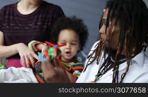 Happy interracial family with parents and curly little toddler son playing with colorful toys in the bedroom at home. Smiling stylish african dad with dreadlocks and mixed race cute child having fun together indoors closeup.