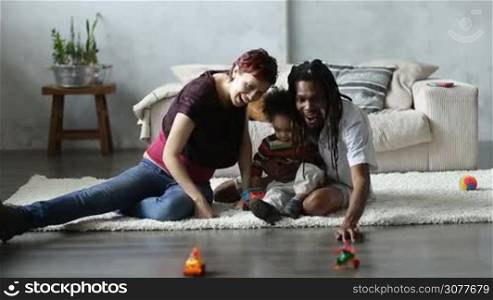 Happy interracial parents sitting on the floor in living room and playing with cute mixed race toddler with colorful toys car. Cheerful mixed race family spending great time together and having fun at home.