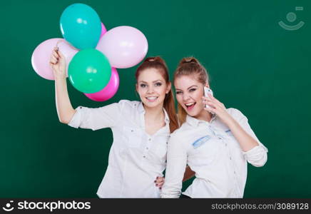 Happy joyful two girls having fun, playing with colorful balloons and talking on mobile phone. Holidays, celebration friendship and technology concept.. Two girls with mobile phone and balloons
