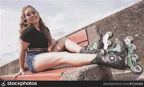 Happy joyful young woman wearing rol≤r skates sitting on bench enjoying herself. Fema≤being sporty having fun during∑mer time.. Young woman riding rol≤r skates