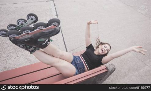 Happy joyful young woman wearing roller skates lying on bench with raised legs. Bizarre high angle. Crazy woman on bench wearing roller skates