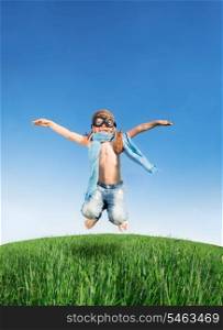 Happy kid dressed as a pilot jumping in green field against blue sky. Summer vacation concept
