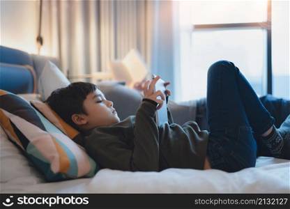Happy Kid lying in bed holding tablet watching cartoon and chatting with friends on digital pad, Cute young boy playing games online on internet, Child relaxing at home with family