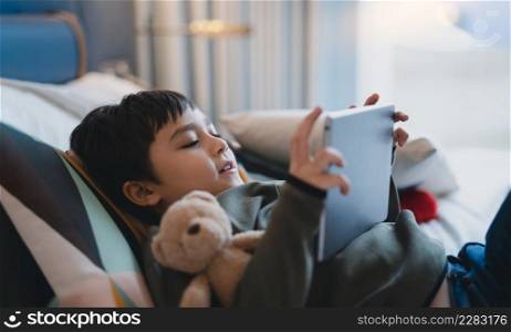 Happy Kid lying in bed holding tablet watching cartoon and chatting with friends on digital pad, Cute young boy playing games online on internet, Child relaxing at home with family