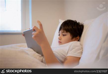 Happy Kid lying in bed holding tablet watching cartoon and chatting with friends on digital pad, Cute young boy playing games online on internet, Child relaxing in the morning before go to school