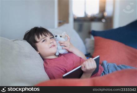 Happy Kid sitting on sofa playing with dog toy and watching cartoons or playing games on tablet, Child boy relaxing after learning online on internet,Home schooling or Online education concept