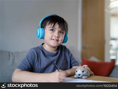 Happy kid wearing headphones listening to music while drawing on paper, Indoors portrait Cute Young boy looking up with smiling face, Positive child enjoy creative activity at home on weekend.