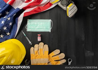 Happy Labor Day during breakout coronavirus. Several constructor work tools, labor COVID-19 vaccine, medical face mask protective and American flag with copy space on black wood background