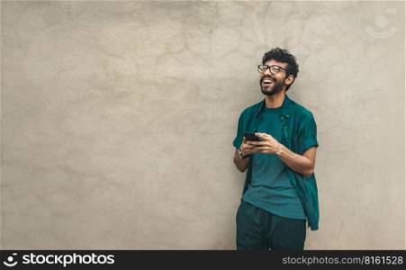 Happy latin man with his cell phone leaning against a wall with copy space, Person smiling phone with space for text. Happy handsome guy leaning against the wall using his cell phone